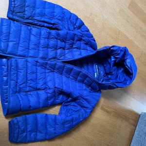 Blå dunjacka från Everest - Säljer en blå tunnare dunjacka från Everest med dragkedja och huva. Jackan är quiltad och har långa ärmar, perfekt för kyligare dagar. Den är lätt och smidig att bära med sig.