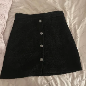 Svart kjol med knappar - Snygg svart kjol med dekorativa knappar framtill. Kjolen har en klassisk A-linjeform och är perfekt för en stilren look. Passar bra till både vardag och festliga tillfällen.