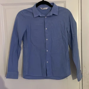 Blå skjorta från H&M - Snygg blå skjorta från H&M med klassisk krage och knappar framtill. Perfekt för en stilren look. Skjortan har långa ärmar och är tillverkad i ett bekvämt material. Storlek 152