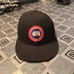 Svart keps från Canada Goose - Snygg svart keps från Canada Goose med ett broderat märke framtill. Kepsen har en justerbar rem baktill för perfekt passform. Perfekt för en stilren look.