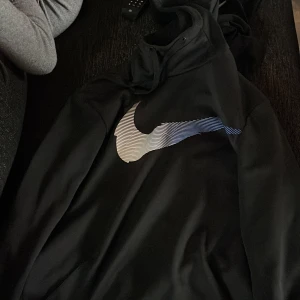 Svart hoodie från Nike - Säljer en svart hoodie från Nike med det klassiska swoosh-loggan i vitt och blått på bröstet. Perfekt för en avslappnad stil.