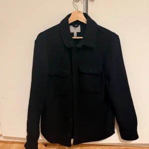 Svart Overshirt från H&M  - Snygg svart jacka från H&M i en blandning av ull, akryl och polyester. Jackan har långa ärmar och knappar framtill. Perfekt för en stilren look. Inga fel på det vad jag vet.