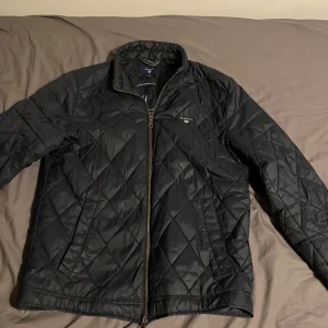 Svart quiltad jacka från GANT - Snygg svart quiltad jacka från GANT med dragkedja framtill. Perfekt för kyligare dagar med sin stilrena design och bekväma passform.  Knappt använd och pris kan diskuteras