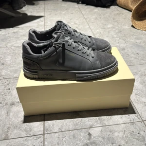 Grå sneakers från Axel Arigato, modell Atlas  - Snygga grå sneakers från Axel Arigato med stilren design. Skorna har snörning och en kombination av skinn och mocka i materialet. Perfekta för en avslappnad och trendig look.