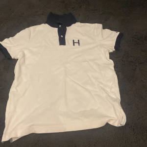Vit pikétröja från Tommy Hilfiger - Snygg vit pikétröja från Tommy Hilfiger med mörkblå krage och ärmslut. Tröjan har en broderad bokstav 'H' på bröstet och knappar vid halsen. Perfekt för en stilren look!