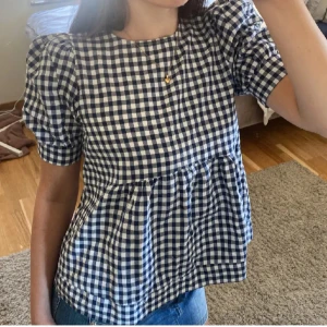 Topp volanger  - Charmig rutig blus i blått och vitt med puffiga kortärmar och rund hals. Perfekt för en avslappnad och stilren look. Blusen har en luftig passform och är lätt att matcha med jeans eller kjol. Priset får alltid att diskuteras 💗