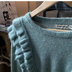 Blå stickad tröja med volanger från Carin Wester - Säljer en mysig grön tröja från Carin Wester med volanger längs ärmarna och framsidan. Tröjan har en rund halsringning och är långärmad. Perfekt för att ge lite extra stil till din garderob!