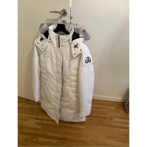 Vit dunjacka från Moose Knuckles - Säljer en snygg vit dunjacka från Moose Knuckles med pälskantad huva och dragkedja. Jackan har ett stilrent märke på ärmen och är perfekt för kalla dagar. Den är lång och håller dig varm och bekväm.