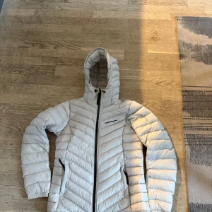 Vit dunjacka från Peak Performance  - Säljer en snygg vit dunjacka från Helly Hansen. Jackan har en dragkedja framtill och praktiska fickor med dragkedjor. Den är långärmad och har en huva för extra värme. Perfekt för kyliga dagar!