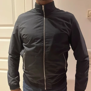 Svart bomberjacka med dragkedja - Snygg svart bomberjacka med dragkedja framtill och på fickorna. Jackan har en stilren design med normal passform och långa ärmar. Perfekt för en cool och avslappnad look.