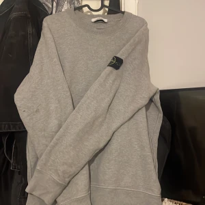 Grå sweatshirt från Stone Island - Säljer en stilren grå sweatshirt från Stone Island. Tröjan har en klassisk rund halsringning och en diskret logga på ärmen. Perfekt för en avslappnad look.