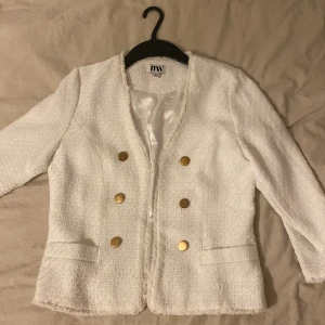 Vit kavaj från Make Way - En lite kortare vit kavaj från Make Way med guldknappar och en stilren design. Kavajen har en texturerad yta och två fickor framtill. 