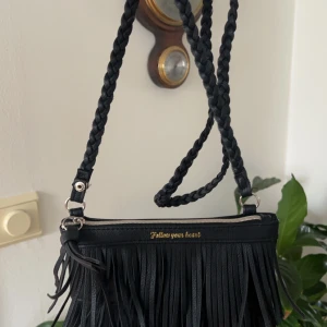 Svart axelväska med fransar - Snygg svart axelväska med flätad axelrem och coola fransdetaljer. Väskan har en dragkedja upptill och texten 'Follow your heart' i guld. Perfekt för en dag på stan eller en kväll ute. 🖤