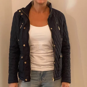 Svart quiltad jacka från Tindra - Säljer en snygg svart quiltad jacka från Tindra. Den har en dragkedja och knappar framtill, samt praktiska fickor med dragkedjor. Perfekt för höst och vår! Jackan är i nyskick och har en normal passform. Perfekt för dig som vill ha en stilren look.