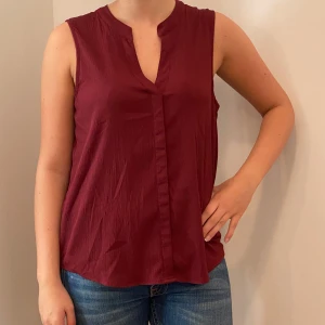 Vinröd ärmlös blus - Säljer en stilren vinröd ärmlös blus i ett lätt och luftigt material. Perfekt för både vardag och festliga tillfällen. Blusen har en v-ringad design och en lös passform som ger en bekväm känsla. Passar perfekt till jeans eller kjol!