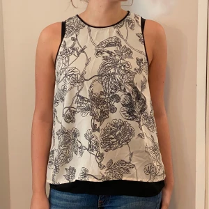 Blommig ärmlös topp från Vero Moda - Säljer en superfin ärmlös topp från Vero Moda i storlek S. Den är vit med ett svart blommigt mönster och har en snygg öppen ryggdetalj. Perfekt för sommardagar eller en avslappnad utekväll. Toppen är i bra skick och väldigt bekväm att bära.