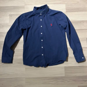 Blå skjorta från Ralph Lauren - Snygg blå skjorta från Ralph Lauren i klassisk passform.  Storlek M men krymt lite så sitter mer som S. Skjortan har långa ärmar och knappar framtill. Den är dekorerad med Ralph Laurens ikoniska röda broderade logotyp på bröstet. Perfekt för en stilren look. Skick 9/10 bara använd lite annars i jätte bra skick. Tveka inte på att höra av dig!