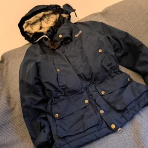 Blå jacka från 8848 Altitude (barn) - Säljer en blå jacka från 8848 Altitude med huva och flera praktiska fickor. Jackan har knappar och dragsko i midjan för justerbar passform. Perfekt för kyligare väder.