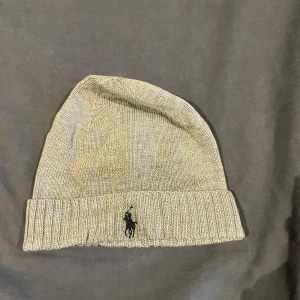 Beige stickad mössa från Polo Ralph Lauren - Snygg beige stickad mössa från Polo Ralph Lauren med broderad logotyp framtill. Perfekt för kyliga dagar med sin mjuka och bekväma passform.
