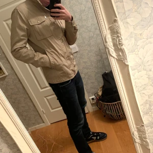 J.lindberg Beige jacka med dragkedja och knappar - Snygg beige jacka med både dragkedja och knappar framtill. Jackan har långa ärmar och flera praktiska fickor. Perfekt för en stilren look.