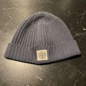 Blå stickad mössa från Stone Island junior - Snygg blå stickad mössa från Stone Island junior med ribbad design och ett broderat märke framtill. Perfekt för att hålla värmen under kyliga dagar. Köpt på NK. Något blekt.