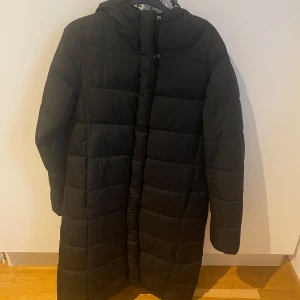 Svart lång dunjacka - Säljer en svart lång dunjacka med huva. Jackan har en quiltad design och stängs med dragkedja. Perfekt för kalla dagar.