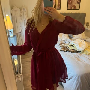 Vinröd klänning med knytband - Säljer en elegant vinröd klänning med knytband i midjan. Klänningen har långa ärmar och en omlott-design som ger en fin passform. Perfekt för höst och vinter med sin djupa färgton. Passar bra till både vardag och festliga tillfällen.