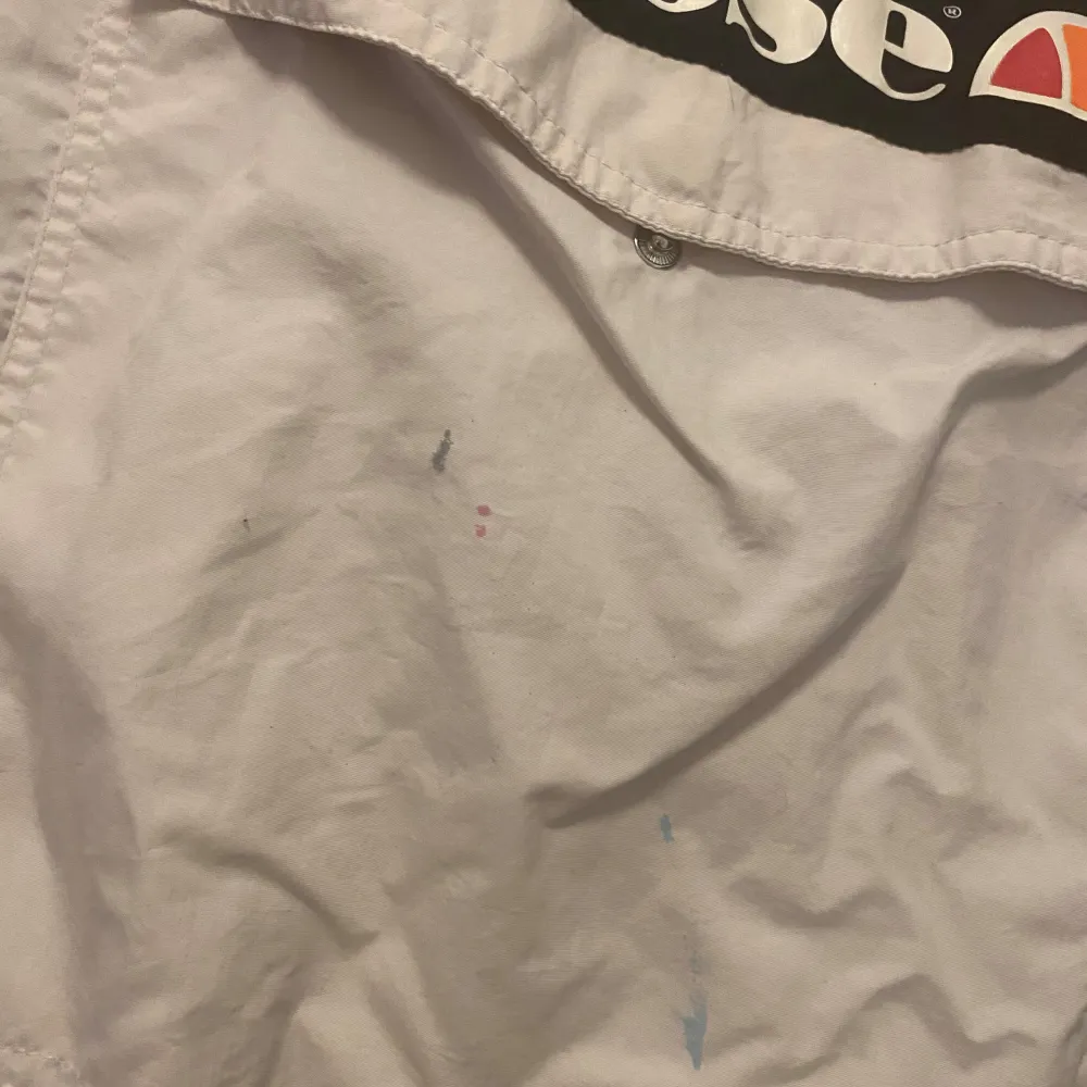 Säljer en vit vindjacka från Ellesse med huva och dragkedja. Jackan har ett stort Ellesse-tryck på framsidan och svarta detaljer längs ärmarna. Perfekt för vår och sommar. Små färgfläckar finns på framsida och ena är en. Se bilder. 🌀. Takit.