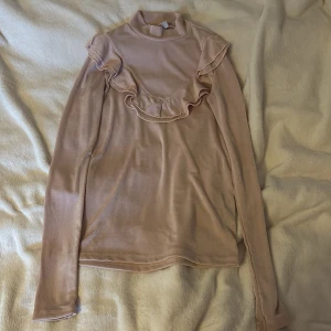 Ljus rosa  långärmad topp från Gina Tricot - Säljer en stilren beige långärmad topp från Gina Tricot med volangdetaljer framtill. Perfekt för en elegant look. Toppen är i ett mjukt material och har en hög krage. Färgen är lite glittrig i sig 