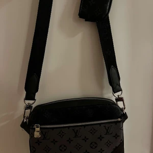 Svart axelväska med monogram - Snygg svart axelväska med klassiskt monogrammönster och justerbar axelrem. Väskan har en praktisk dragkedja och detaljer i metall. Perfekt för att bära dina nödvändigheter med stil.Kom privat för mer info