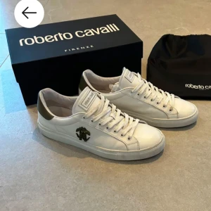 Vita sneakers från Roberto Cavalli - Snygga vita sneakers från Roberto Cavalli med snörning och en stilren design. Skorna har en diskret logga på sidan och en kontrasterande detalj i mörkare färg vid hälen. Perfekta för en trendig look. Storlek 45 och väldigt fräscha.