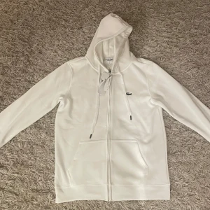 Vit hoodie från Lacoste - Säljer en HELT NY stilren vit hoodie från Lacoste med dragkedja och huva. Tröjan har långa ärmar och en liten krokodil-logga på bröstet. Perfekt för en avslappnad look. Tröjan är S men passar även XS