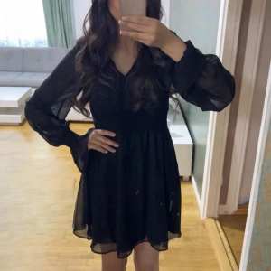 Svart klänning med v-ringning - Snygg svart klänning med v-ringning och långa, transparenta ärmar. Den har en smockdetalj i midjan som ger en fin passform. Perfekt för en stilren look.