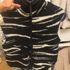 Svart och vit fleeceväst från H&M - Säljer en cool svart och vit fleeceväst från H&M med zebramönster. Västen har en dragkedja framtill och är perfekt för att hålla sig varm och stilren. Passar bra för höst och vinter.