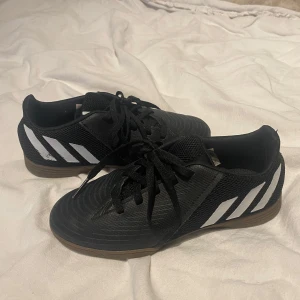 Svarta fotbollsskor från Adidas - Säljer ett par svarta fotbollsskor från Adidas med vita ränder på sidorna. Skorna har snörning och en brun sula. Perfekta för inomhusfotboll och har en stilren design.