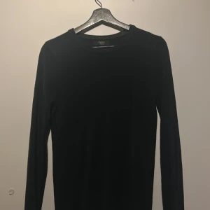 Svart långärmad tröja från Next - Säljer en stilren svart långärmad tröja från Next. Perfekt för kyligare dagar med sin enkla design och bekväma passform. Tröjan har en rund halsringning och är tillverkad i ett mjukt material.