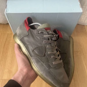  Philippe Model trainers - Snygga grå sneakers från Philippe Model med röda detaljer. Skorna har en klassisk snörning och en robust sula. Perfekta för en stilren look. Väldigt bra skick samt box fås med