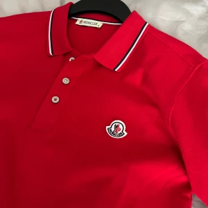 Röd pikétröja från Moncler - Snygg röd pikétröja från Moncler med klassisk krage och tre knappar framtill. Tröjan har korta ärmar och Monclers logga broderad på bröstet. Perfekt för en stilren look. F A K E men man märker inte av.Är inte använd nån gång hör av vid frågor!