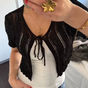 Svart spetsbolero - Säljer en elegant svart spetsbolero med korta ärmar och knytning framtill. Perfekt för att lägga till en touch av stil till din outfit. Passar bra över en enkel topp eller klänning.