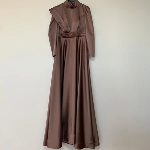 Brun långklänning i satin - Elegant brun långklänning i satin med långa ärmar och hög hals. Klänningen har en unik design med draperade detaljer över axlarna och en markerad midja som ger en smickrande silhuett.