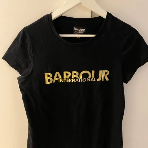 Svart t-shirt från Barbour - Snygg svart t-shirt från Barbour med guldig logga på bröstet. Perfekt för en stilren look. Kortärmad och i mjukt material för bekväm passform.