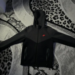 Nike tech - Svart och grå Nike tech zip tröja, tröjan har full zip och Nike märket är i färgen röd, storleken är M men skulle nog säga mer som S. nypris var runt 2500, pris kan diskuteras. 