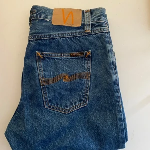Nudie Jeans - Säljer ett par klassiska blå jeans från Nudie Jeans. De har en snygg straight passform och är tillverkade i slitstarkt denim. Byxorna har en midwaist design och är perfekta för en avslappnad stil. Detaljer som knappar och Nudie-loggan på bakfickan ger en cool touch. Stick 9/10