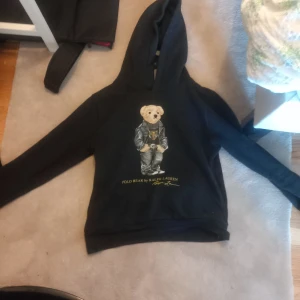 Svart hoodie från Ralph Lauren med björnmotiv - Säljer en svart hoodie från Ralph Lauren med ett ikoniskt björnmotiv på framsidan. Tröjan har en klassisk huva och långa ärmar, perfekt för en avslappnad stil. Märket är välkänt för sin kvalitet och stil. Passar perfekt för kyligare dagar. Öppen för byten