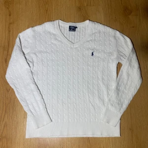 Vit stickad Ralph Lauren - Säljer en stilren vit stickad tröja från Ralph Lauren med klassiskt kabelstickat mönster. Tröjan har en v-ringad hals och långa ärmar. Den ikoniska loggan är broderad på bröstet i marinblått. Perfekt för en elegant och avslappnad stil.   Storlek L men passar dig om du är 150-164 så S eller Xs