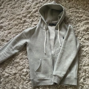 Grå hoodie från Ralph Lauren - Säljer en stilren grå hoodie från Ralph Lauren med dragkedja och justerbar huva. Tröjan har en broderad logga på bröstet. Oandvänd från Zalando. Pris diskuterbart
