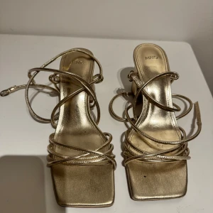 Guldiga klackskor från Mango - Klackskor från Mango med smala remmar som knyts runt ankeln. Perfekta för studentbal/examensfirande😍Använde dessa på min studentbal och det är det ända tillfället dom har använts. Slutsålda och nypris 529kr.