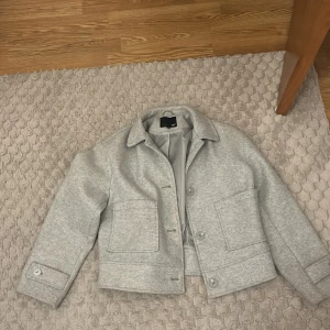 Grå kappa från 157 - Säljer en stilren grå kappa från 157 i storlek M. Jackan har en klassisk krage och knappar framtill. Den är perfekt för kyligare dagar och har praktiska fickor på framsidan.