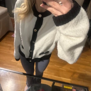 Vit och svart kofta med knappar - Säljer en mysig vit kofta med svarta detaljer och knappar. Koftan har långa ärmar och en klassisk design med ribbade kanter. Perfekt för att hålla sig varm och stilren.