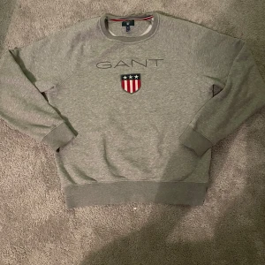 Grå sweatshirt från GANT - Säljer en stilren grå sweatshirt från GANT med ett tryckt emblem på bröstet. Tröjan har långa ärmar och rund halsringning. Perfekt för en avslappnad look.